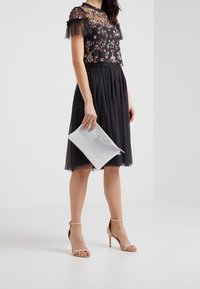 Musta mekko, jossa on kukkakirjontaa, läpikuultavat pitsihihat ja teksturoitu hame. Yhdistetty hopeiseen clutch-laukkuun ja nude-hihnakenkiin.