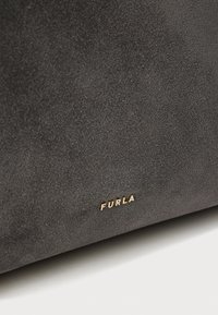 Surface en daim gris foncé avec de petites lettres en métal doré formant le mot "FURLA" près du bord inférieur.