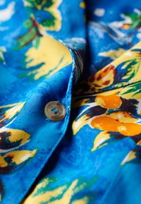 Tejido azul con un estampado de frutas tropicales que incluye naranjas y plátanos, complementado por un botón de color claro en la zona del cuello.