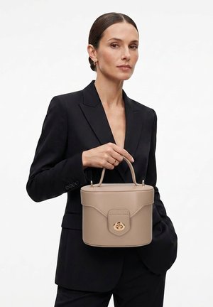 Vrouw in zwart pak die een beige gestructureerde handtas met bovenhandvat en gouden gesp vasthoudt tegen een effen achtergrond.