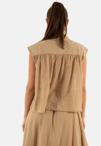 Top sin mangas en beige con un corte holgado, detalle de pliegues en la parte posterior y textura suave, llevado sobre una falda a juego, mostrado desde atrás.