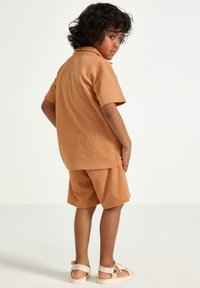 Terry stof sæt i lysebrun, bestående af en kortærmet skjorte med krave og matchende shorts, båret med beige sandaler.