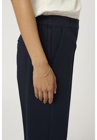 Bracelet en argent au poignet, avec un design délicat. La personne porte un pantalon marine et un haut de couleur claire, sur un fond uni.