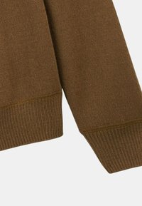 Manche de pull en tricot marron avec poignet côtelé sur fond gris clair, montrant la texture et les détails de couture.