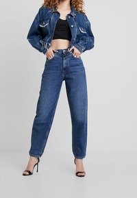 Farkkutakki kuluneilla yksityiskohdilla, musta lyhyt toppi ja korkeavyötäröiset siniset farkut. Jalkineina mustat remmikorolliset korkokengät.