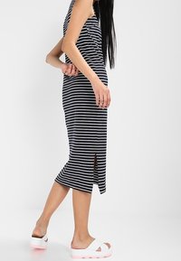 Abito midi a righe, senza maniche, in blu scuro e bianco, con un'apertura laterale. Indossato con sandali bianchi che mostrano una suola rosa.