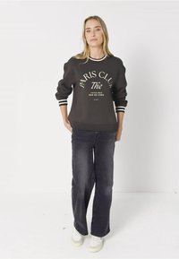 Sweat à capuche noir avec le texte "PARIS CLUB" en blanc, poignets rayés, associé à un jean large bleu foncé et des baskets blanches, se tenant contre un fond neutre.