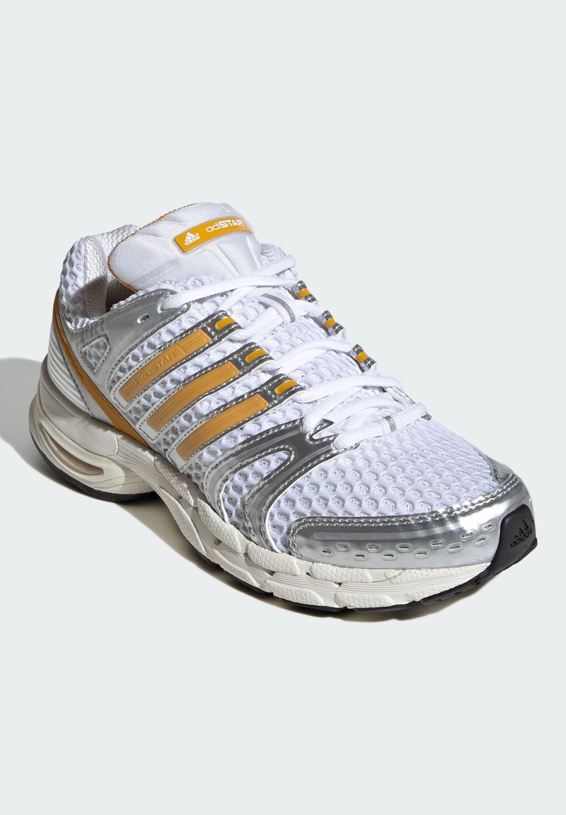 Chaussure de course Adidas blanche et argentée avec des accents jaunes, empeigne en mesh, semelle rembourrée et fermeture à lacets sur un fond clair uni.