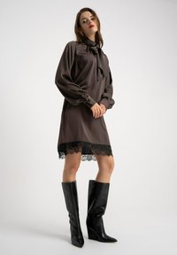 Brązowa suknia z satyny z długimi bufiastymi rękawami i kokardą przy szyi; posiada czarną koronkową lamówkę u dołu. Połączona z wysokimi czarnymi botkami.