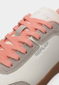 Gros plan sur une basket blanche et grise avec des lacets corail, des détails en daim gris, et le logo "REPLAY" sur le côté et la languette.