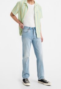 Camisa de manga corta con botones y rayas multicolores en verde, amarillo y azul; camiseta blanca sin mangas debajo; vaqueros azul claro; zapatillas negras.