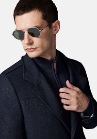 Męski granatowy teksturowany płaszcz z zamkiem błyskawicznym, pod którym znajduje się ciemny sweter. Okrągłe, metalowe okulary przeciwsłoneczne z zielonymi soczewkami.