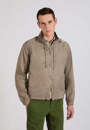 Mężczyzna w beżowej kurtce zapinanej na zamek z troczkami i oliwkowozielonych spodniach, stojący z jedną ręką w kieszeni na tle jednolitego tła.