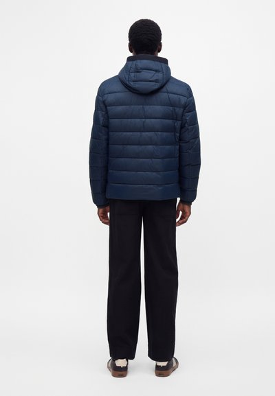 Adulte portant une veste matelassée bleu marine avec capuche, pantalon noir et baskets décontractées, debout de dos sur un fond blanc uni.