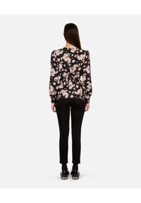 Vrouw met lang donker haar, gekleed in een zwarte bloempatronen lange mouw top, zwarte slim fit broek en zwarte schoenen, staande met haar rug naar de kijker.