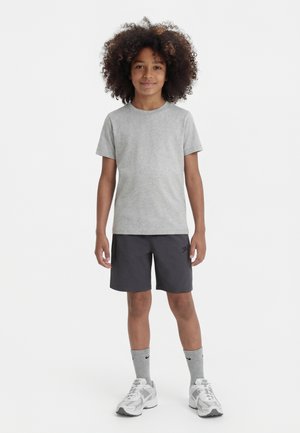 Niño con cabello rizado que lleva una camiseta gris, pantalones cortos negros, calcetas grises y zapatillas blancas, de pie contra un fondo blanco.