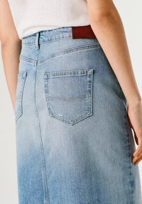 Prim-plan cu o fustă din denim albastru deschis cu buzunare în spate și o cămașă albă purtate de o persoană care stă cu mâna relaxată pe lângă corp.