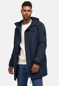 Veste imperméable bleu marine avec capuche, fermeture éclair à l'avant, plusieurs poches, poignets texturés et un détail en patch sur la manche. Doublure intérieure visible.