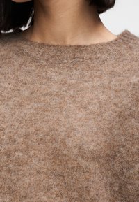 Καφέ πουλόβερ με απαλή, χνουδωτή υφή, με στρογγυλή λαιμόκοψη και ριμπ λεπτομέρειες. Το ύφασμα παρουσιάζει διακριτικές παραλλαγές στο χρώμα.