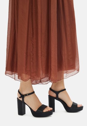 Czarne sandały na wysokim obcasie o fakturowanej powierzchni, z odkrytymi palcami, paskiem wokół kostki i masywnym obcasem, zestawione z lekką, przezroczystą brązową spódnicą.