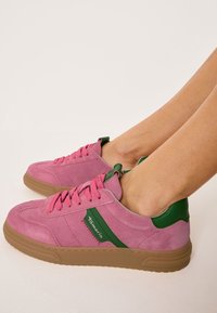 Růžové semišové tenisky se zelenými detaily a růžovými tkaničkami. Mají gumovou podrážku a hladkou texturu, s nízkým střihem.