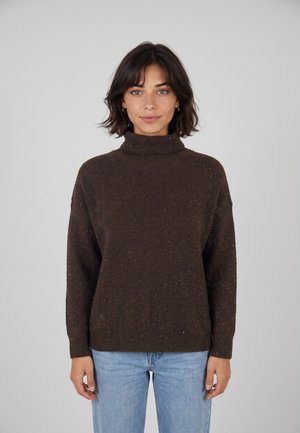 Donna con capelli corti e scuri che indossa un maglione dolcevita marrone scuro e jeans azzurri chiari, in piedi davanti a uno sfondo chiaro e semplice.