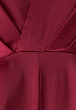 Zbliżenie na głęboki czerwony satynowy materiał z symetrycznymi fałdami i szwami tworzącymi gwiaździsty wzór.