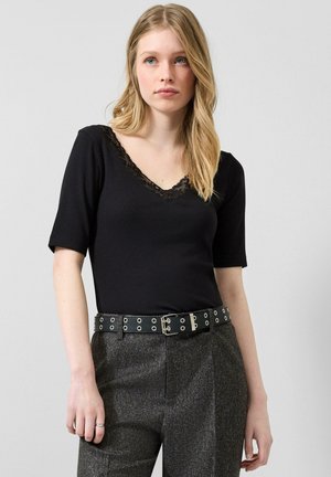 Junge Frau mit langen blonden Haaren, die ein schwarzes, mit Spitze besetztes Oberteil, graue hoch taillierte Hose und einen schwarzen Gürtel mit Ösen trägt, vor einem grauen Hintergrund steht.