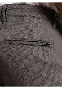 Grijze textuurstofbroek met een achterzak voorzien van een dunne horizontale opening. Een klein zwart label met "GABBA" is zichtbaar.