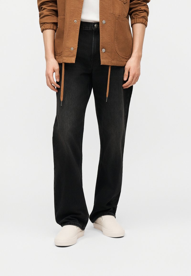 Άτομο που φοράει μαύρο τζιν με φαρδιές παντελόνες, λευκά παπούτσια χωρίς κορδόνια, καφέ μπουφάν με κορδόνια και λευκό πουκάμισο μπροστά σε απλό φόντο.