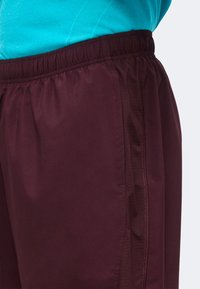 Gros plan d'une personne portant un short bordeaux à taille élastique et une chemise turquoise, montrant les détails de la couture latérale et la texture du tissu.