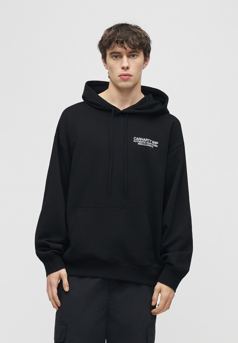 Giovane uomo che indossa una felpa con cappuccio nera Carhartt WIP con logo bianco sul petto, in piedi davanti a uno sfondo grigio chiaro uniforme.