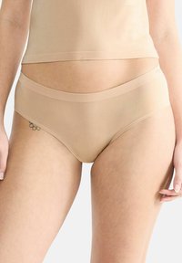 Nahaufnahme einer Person, die beiges nahtloses Unterwäsche und ein passendes Oberteil trägt, mit einem kleinen Blumentattoo, das auf der linken Hüfte sichtbar ist.