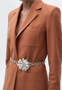 Blazer texturé marron avec grande poche avant, assorti d'une ceinture florale argentée ornée d'accents en chaîne circulaires et d'un détail central en forme de fleur.