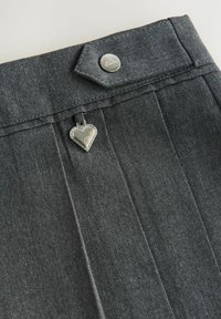 Pantaloni a pieghe grigi con chiusura a bottone, un ciondolo a forma di cuore in metallo e un tessuto testurizzato. Logo del brand in modo discreto sul bottone.