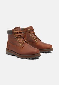 Botas de tobillo de cuero marrón con ojales dorados, cordones marrones, un zipper lateral y una suela de goma resistente. Presenta un logo en el costado.