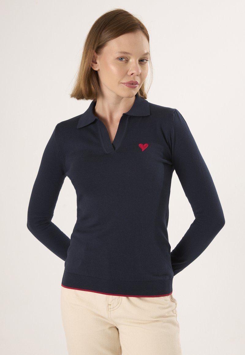 Navy blauw poloshirt met lange mouwen en een rood hart-embleem op de borst, voorzien van een kraag en geribbelde zoom. Crème-kleurige broek.