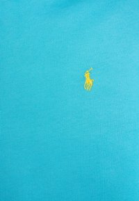 Polo de algodón color turquesa con textura acanalada, que presenta un pequeño logo bordado amarillo de un jinete en el área del pecho izquierdo.