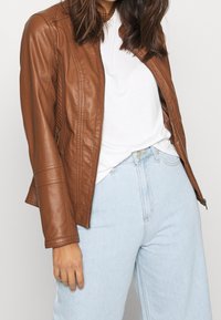 Giacca in pelle marrone con un design aderente, chiusura con zip e dettagli di cuciture testurizzate, abbinata a una maglietta bianca e jeans azzurri chiari.