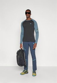 Mann med mørke solbriller, svart og blå treningstøy, holder en svart ryggsekk, står mot en enkel lys bakgrunn.