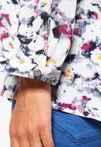 Manche de chemise à motif floral en blanc avec des accents violets, roses et jaunes. Elle comporte un poignet boutonné avec une quincaillerie argentée ornée.