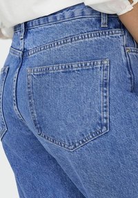 Jasnoniebieskie dżinsy o gładkiej fakturze, wyróżniające się wysokim stanem, tylnymi kieszeniami oraz kontrastowymi detalami w szwach.