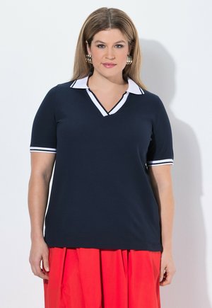 Kobieta z jasnobrązowymi włosami ubrana w granatowy krótki top z białym kołnierzykiem i czerwoną plisowaną spódnicę, stojąca na białym tle.