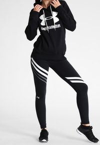 Czarna bluza z kapturem z białym logo Under Armour, połączona z czarnymi legginsami z białymi i szarymi ukośnymi paskami oraz czarnymi butami sportowymi.