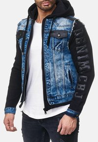 Denim en fleece jack met een blauwe denim romp, zwarte fleece mouwen, ritssluiting en schouderpatches. "DENIM" is op de mouw gestikt.