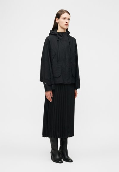 Femme debout portant une veste à capuche noire, une jupe midi plissée noire et des bottes noires à talons montant jusqu'au genou, sur un fond clair uni.