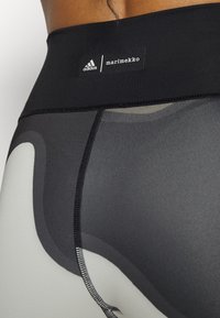 Černé legíny s vysokým pasem, které kombinují matný a lesklý design, zdobené šedými a černými abstraktními vzory. Obsahují loga Adidas a Marimekko.