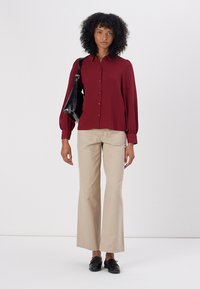 Cămașă roșie cu mânecă lungă și nasturi, cu un finisaj texturat, asociată cu pantaloni bej evazați și pantofi loafers negri, purtând o geantă neagră.