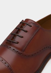 Καφέ δερμάτινο ανδρικό παπούτσι με διακόσμηση brogue και μαύρα κορδόνια, κοντινή λήψη στο πάνω μέρος και το ράψιμο σε λευκό φόντο.