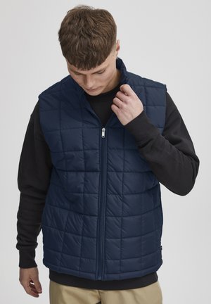 Navy blue padded vest with a quilted pattern and high collar, featuring a front zipper and a relaxed fit, worn over a black long-sleeve shirt.